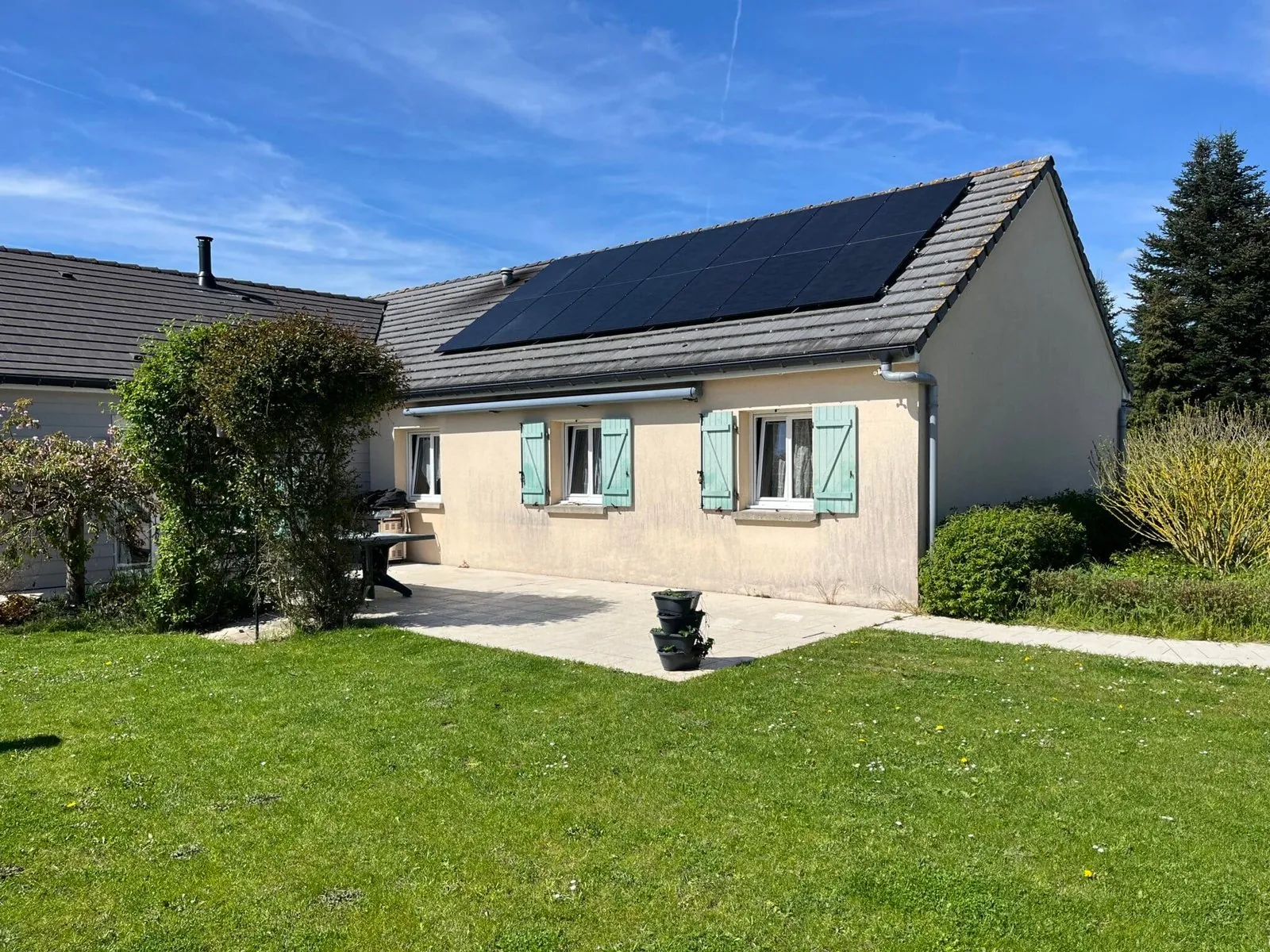 Maison avec des volets verts et panneaux solaires sur le toit, entourée de végétation et sous un ciel bleu clair.
