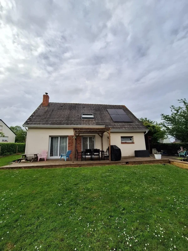 Vue arrière d'une maison avec terrasse, chaises et panneaux solaires sur le toit, sous un ciel nuageux.