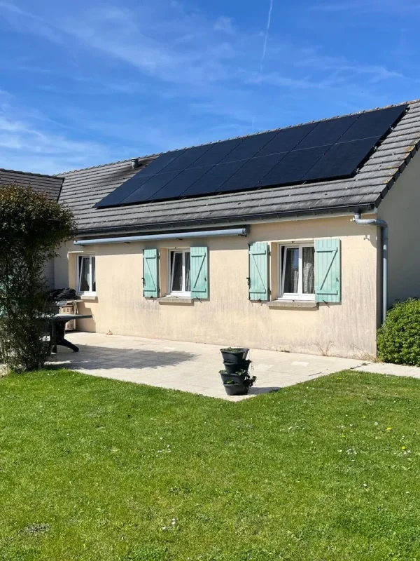 Maison avec des volets verts et panneaux solaires sur le toit, entourée de végétation et sous un ciel bleu clair.